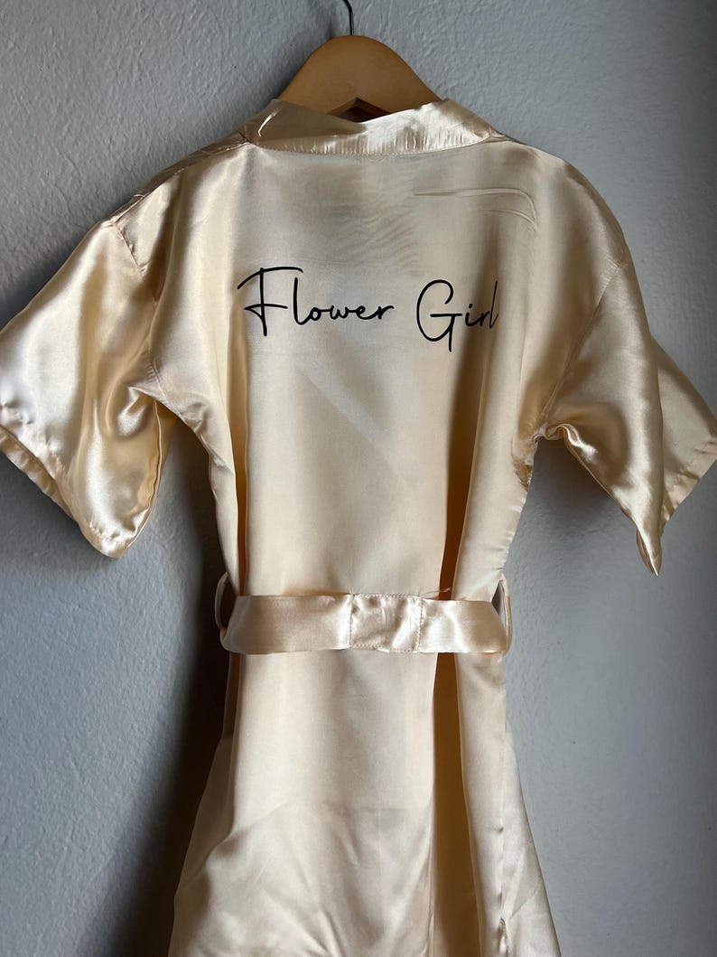 Op de afbeelding: Een cr&egrave;mekleurige satijnen badjas met korte mouwen en een ceintuur. De badjas heeft de woorden "Flower Girl" in het zwart geborduurd op de achterkant.