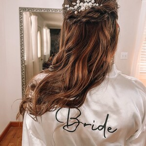 Op de afbeelding: Een witte satijnen badjas met het woord "Bride" geborduurd op de rug. De badjas heeft een haaraccessoire van parels en zilver in het haar.