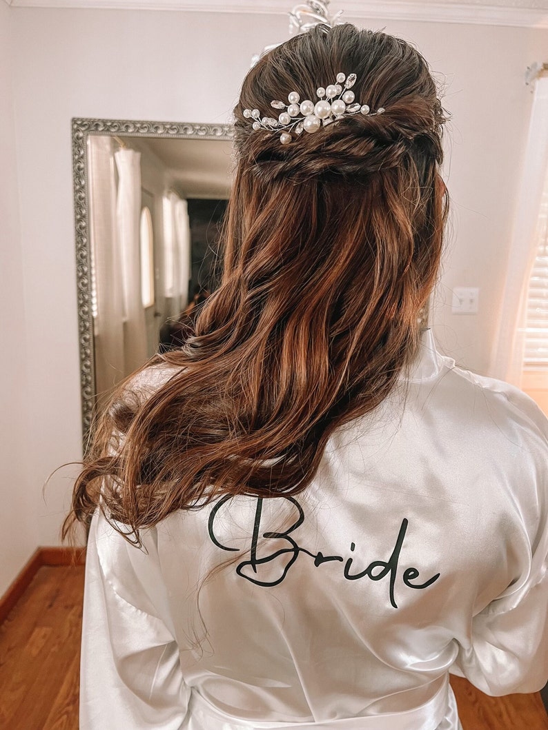 Op de afbeelding: Een witte satijnen badjas met het woord "Bride" geborduurd op de rug. De badjas heeft een haaraccessoire van parels en zilver in het haar.