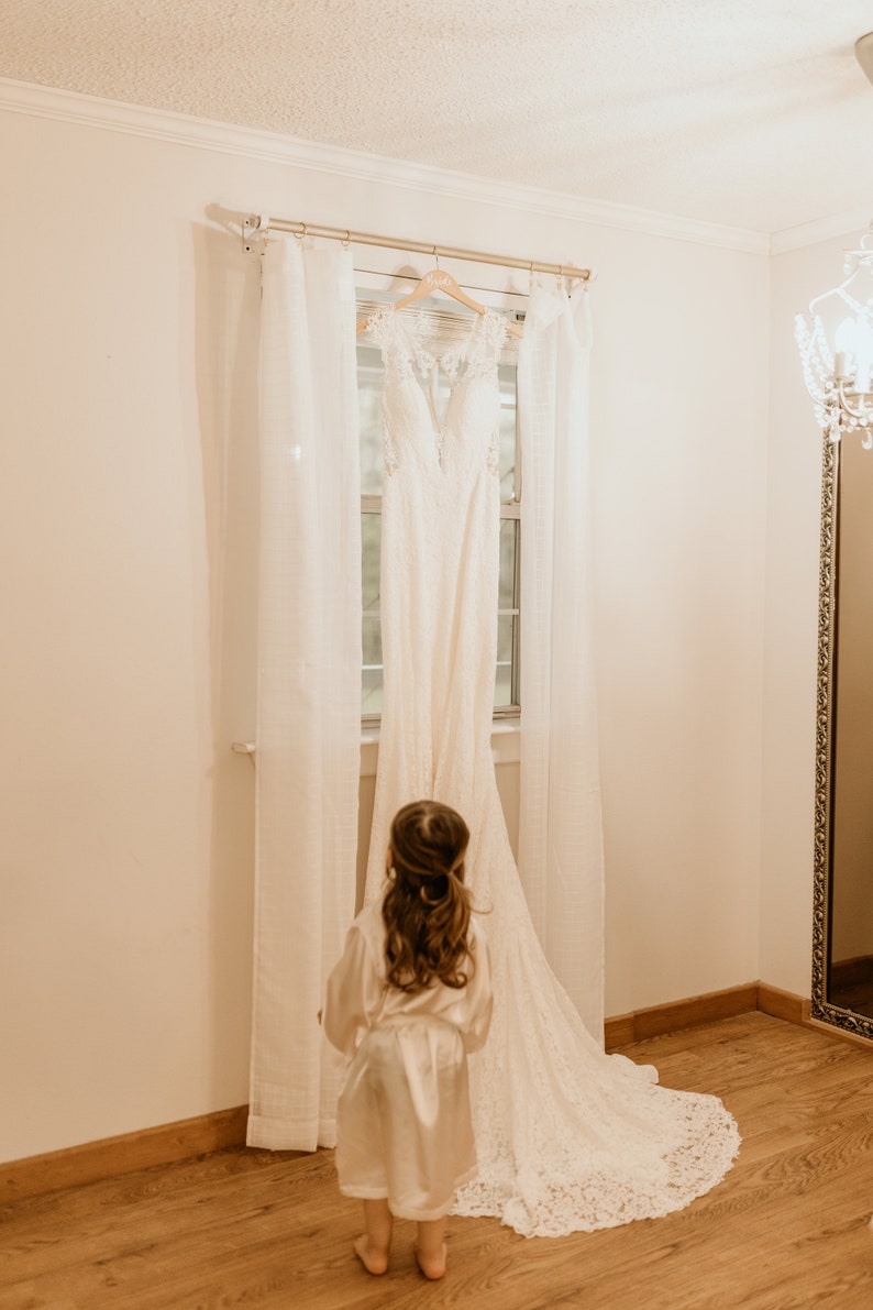 Op de afbeelding: Een jong meisje in een witte satijnen badjas kijkt naar een witte bruidsjurk die aan een hanger hangt voor een raam met witte gordijnen. De jurk heeft een kantbovenstuk en een zwierige rok.