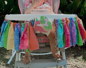 Könnte beinhalten: Ein Regenbogen-Stoffbanner mit Quasten hängt um einen Hochstuhl. Ein kleines Mädchen sitzt im Stuhl und trägt ein rosa Shirt und einen rosa-grünen Dinosaurier-Strampler. Das Banner hat einen rosa Filzdinosaurier mit einem grünen Blatt und der Zahl "1" darauf.