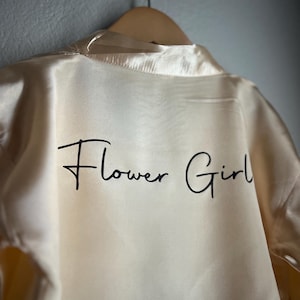 Op de afbeelding: Een cr&egrave;mekleurige satijnen badjas met de tekst "Flower Girl" in zwarte schrijfletters.