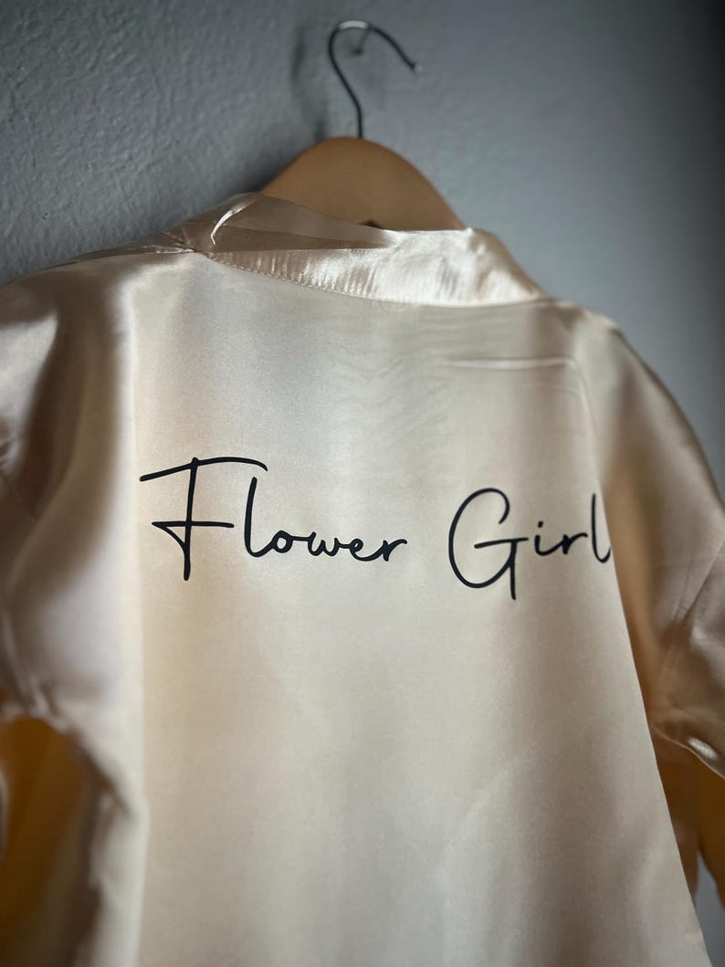 Op de afbeelding: Een cr&egrave;mekleurige satijnen badjas met de tekst "Flower Girl" in zwarte schrijfletters.