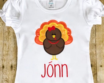 May include: White short-sleeved shirt with a ruffled hem, featuring an embroidered turkey with a red bow and the name "Jõnn" in red.