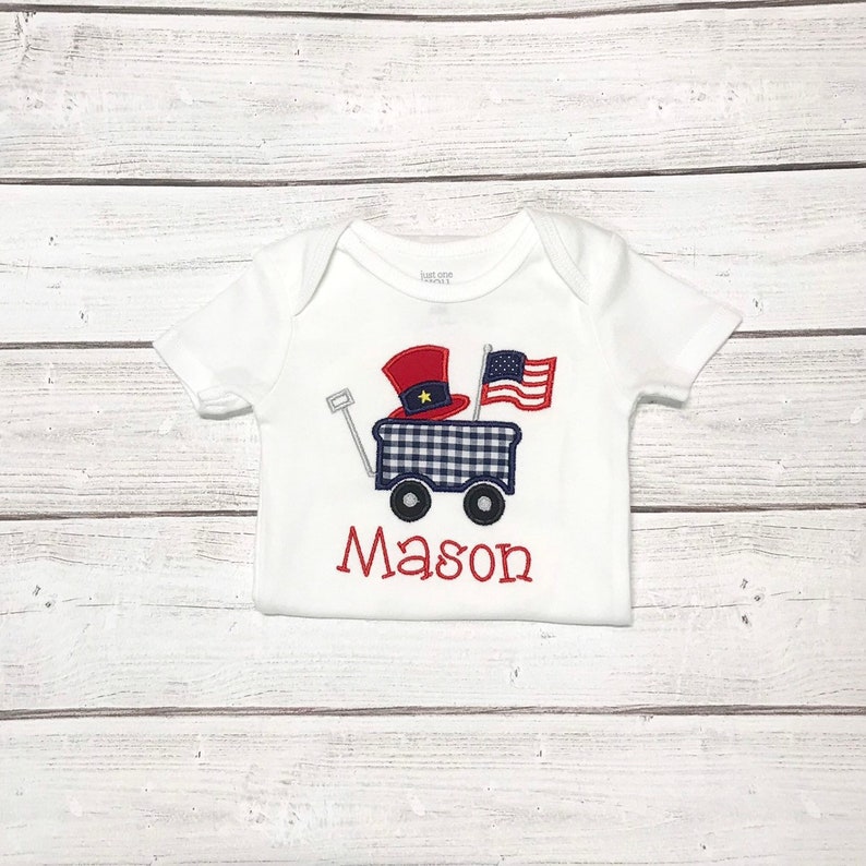 Op de afbeelding: Een witte baby romper met een rood, wit en blauwe wagen met een Amerikaanse vlag en een rood-witte hoed. De naam "Mason" is in rood geborduurd onder de wagen.