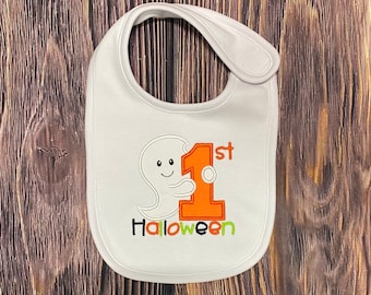 Peut inclure: Bavoir blanc pour bébé avec un fantôme brodé tenant un grand numéro un orange avec le mot "1er" au-dessus et le mot "Halloween" en dessous.