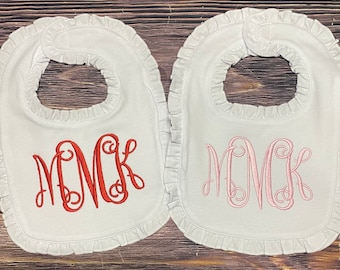 以下が含まれることがあります： フリル付きの白いベビーよだれかけが2枚。1枚は赤い刺繍で「MCK」のモノグラムが施され、もう1枚はピンクの刺繍で「MCK」のモノグラムが施されています。