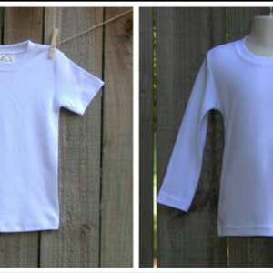 Op de afbeelding: Twee witte T-shirts, een met korte mouwen en een met lange mouwen, hangen aan een houten hek.