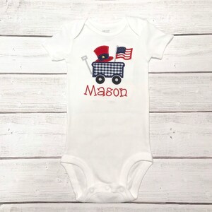 Op de afbeelding: Witte baby romper met een rood, wit en blauwe wagen met een rood en blauwe hoed en een Amerikaanse vlag. De naam "Mason" is geborduurd op de romper.