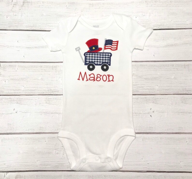 Op de afbeelding: Witte baby romper met een rood, wit en blauwe wagen met een rood en blauwe hoed en een Amerikaanse vlag. De naam "Mason" is geborduurd op de romper.