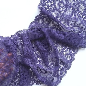 May include: A length of deep purple lace fabric with a floral pattern. The lace is folded and draped, showcasing its intricate design and delicate texture. The fabric is against a white background.