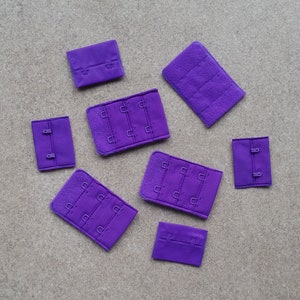Könnte beinhalten: Acht violette Stoff-BH-Haken- und Ösenverschlüsse in verschiedenen Größen.
