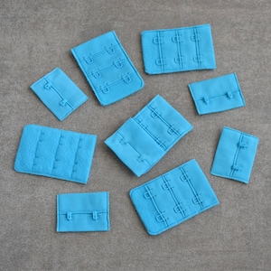 May include: A collection of light blue hook and eye closures, in various sizes, arranged on a gray surface. The closures are made of fabric and feature multiple hook and eye sets, with stitched detailing.