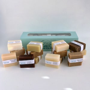 May include: A box of 12 bars of handmade shampoo. The bars are stacked in groups of three and are labeled with different scents, including Apple Cider Vinegar, Cassia, Aloe & Jojoba, and  Bean &  Milk. The box is light blue with a clear window.