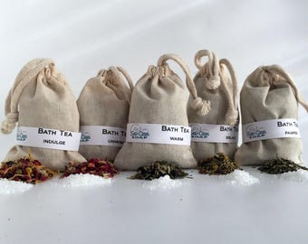May include: Six small white muslin bags filled with bath tea blends. The bags are tied with twine and labeled with the words "Bath Tea" and different scents, including "Indulge", "Unwind", "Warm", "Relax", and "Pamper".