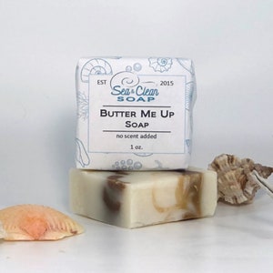 A white bar of soap with a brown swirl design. The soap is labeled 'Butter Me Up Soap' with the words 'Sea & Clean Soap' and 'no scent added' printed on the label. The soap is sitting on a white surface with seashells.