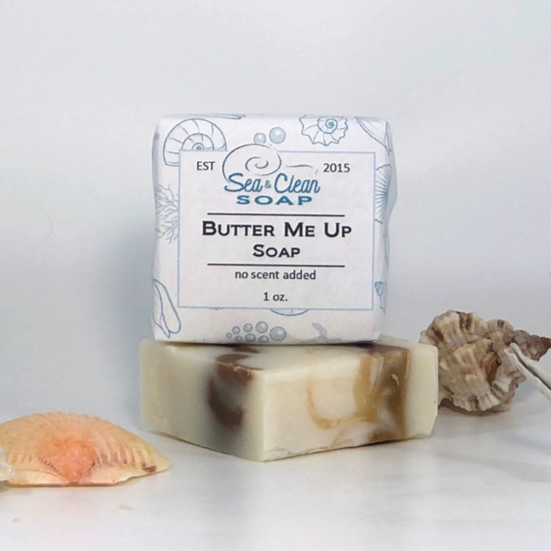 A white bar of soap with a brown swirl design. The soap is labeled 'Butter Me Up Soap' with the words 'Sea & Clean Soap' and 'no scent added' printed on the label. The soap is sitting on a white surface with seashells.