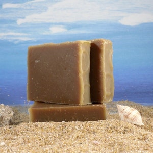May include: Three bars of brown soap stacked on top of each other on a sandy beach with a seashell in the foreground.