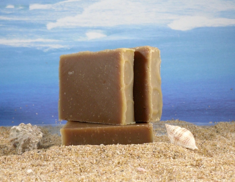 May include: Three bars of brown soap stacked on top of each other on a sandy beach with a seashell in the foreground.