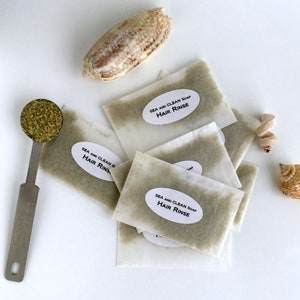 Może przedstawiać: Stos małych białych papierowych saszetek wypełnionych zielonym proszkiem. Każda saszetka ma białą etykietę z napisem "Sea and Clean Soap Hair Rinse".