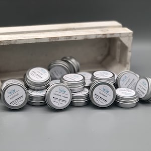 May include: A collection of small silver tins filled with body powder. The tins are labeled with different scents, including lavender, coconut cream, and patchouli. The tins are arranged in a pile on a gray surface.