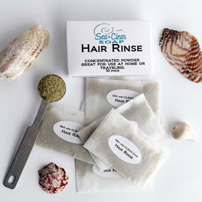 A white box with the text 'Sea & Clean Soap Hair Rinse' on it. The box is next to a pile of white paper packets with the text 'Sea and Clean Soap Hair Rinse' on them. The packets are filled with a green powder. The image also includes seashells and a measuring spoon.