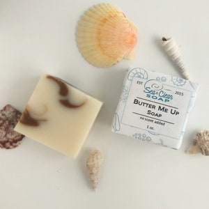 A white bar of soap with brown swirls.  Another soap bar wrapped in paper. The label has the text 'Sea & Clean SOAP' and 'BUTTER ME UP SOAP' with the text 'no scent added' and '1 oz.' The box is surrounded by seashells.