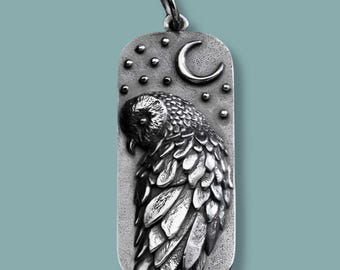 May include: A silver pendant featuring a detailed owl design. The pendant has a rectangular shape with a textured surface. The owl is depicted with intricate feather details, a crescent moon, and stars.