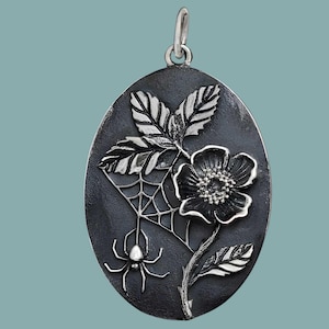 Op de afbeelding: Ovale hanger met een donkere achtergrond, met een gedetailleerd zilveren ontwerp. Het ontwerp omvat een bloem, bladeren, een spinnenweb en een spin. De hanger heeft een zilveren lus aan de bovenkant voor een ketting.