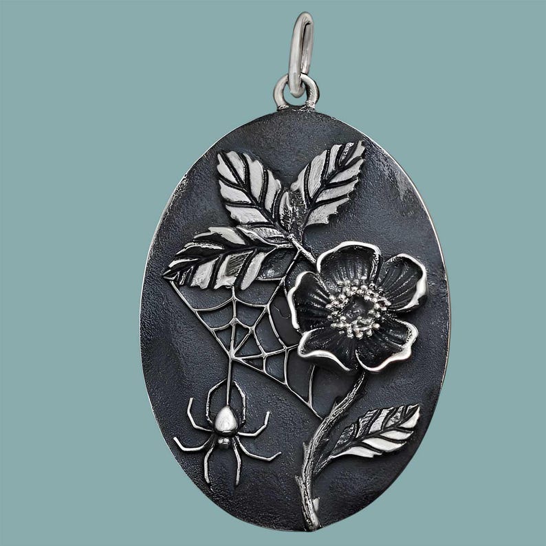 Op de afbeelding: Ovale hanger met een donkere achtergrond, met een gedetailleerd zilveren ontwerp. Het ontwerp omvat een bloem, bladeren, een spinnenweb en een spin. De hanger heeft een zilveren lus aan de bovenkant voor een ketting.