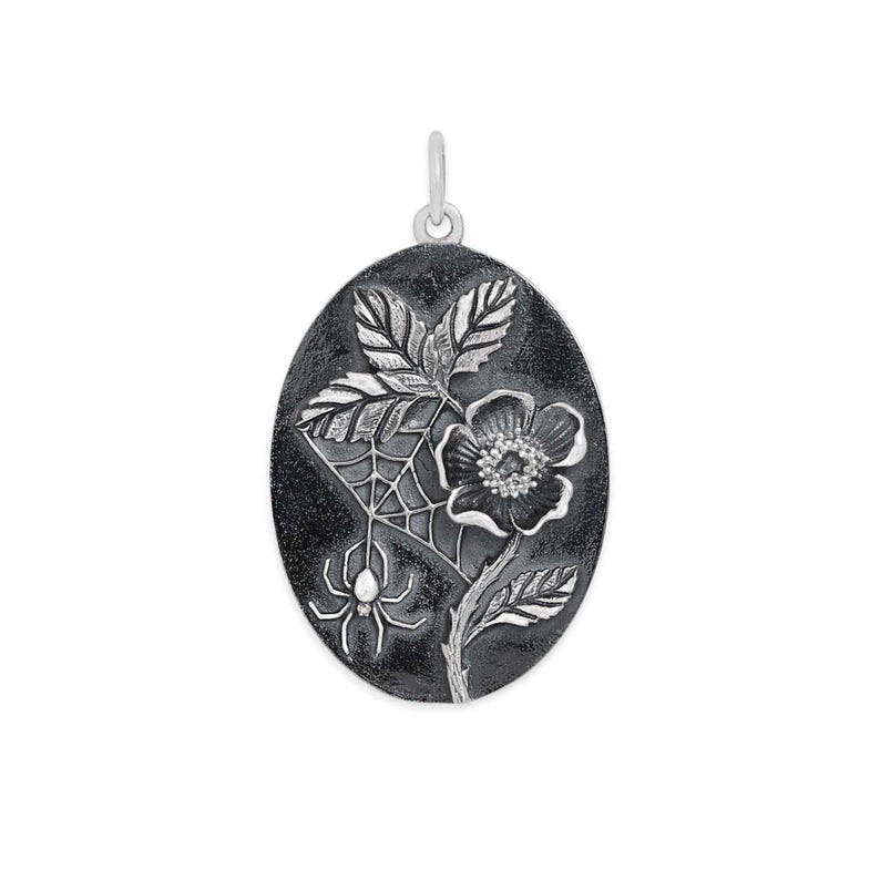 Op de afbeelding: Zilverkleurige ovale hanger met een bloemen- en spinnenwebontwerp. De hanger heeft een gedetailleerde bloem, bladeren en een spinnenweb met een spin. Het heeft een oogje om aan een ketting te bevestigen.