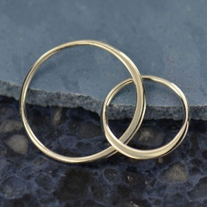 May include: Two gold-colored, circular hoop earrings are interlocked, set against a dark blue, textured surface. The earrings are simple in design, with a smooth, polished finish, and are likely made of metal.