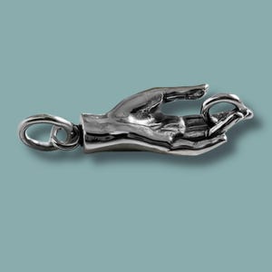 Puede incluir: Un charm de color plateado con forma de mano sosteniendo un anillo. El charm tiene un diseño detallado y realista e incluye un lazo para sujetarlo a un collar o pulsera. El fondo es de color verde azulado.