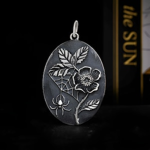 Op de afbeelding: Ovale zilveren hanger met een gedetailleerd bloemmotief, spin en web. De hanger toont een bloem, bladeren en een spinnenweb met een spin. Het heeft een lus aan de bovenkant voor een ketting.