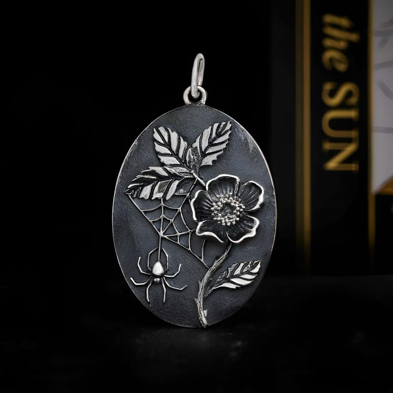 Op de afbeelding: Ovale zilveren hanger met een gedetailleerd bloemmotief, spin en web. De hanger toont een bloem, bladeren en een spinnenweb met een spin. Het heeft een lus aan de bovenkant voor een ketting.