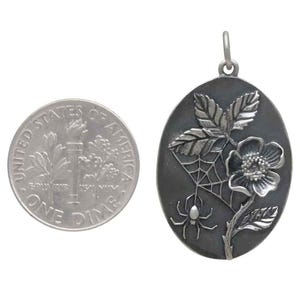 Op de afbeelding: Ovale zilveren hanger met een bloemen- en spinnenwebontwerp. Een Amerikaanse dime ligt naast de hanger ter vergelijking van de grootte. De hanger heeft een bloem, bladeren, een spinnenweb en een spin.