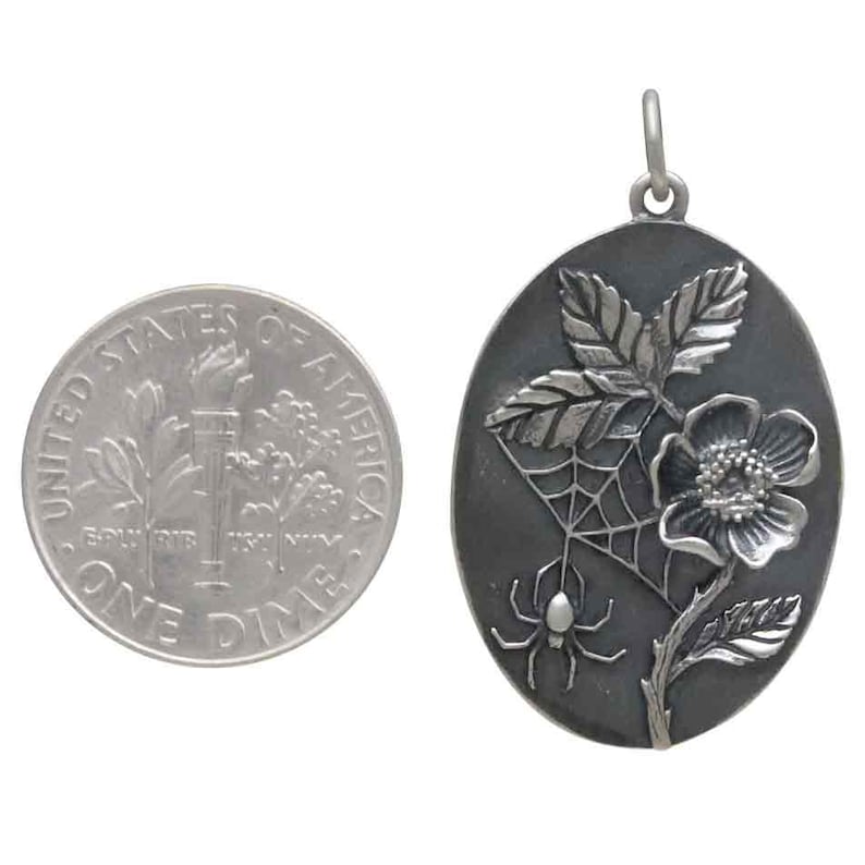 Op de afbeelding: Ovale zilveren hanger met een bloemen- en spinnenwebontwerp. Een Amerikaanse dime ligt naast de hanger ter vergelijking van de grootte. De hanger heeft een bloem, bladeren, een spinnenweb en een spin.