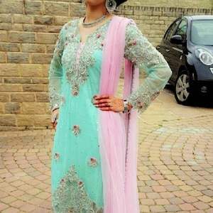 May include: A woman wearing a light blue and pink embroidered dress with a pink dupatta. She is standing on a brick pathway.