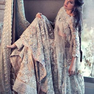May include: An intricately designed, light-colored formal gown with elaborate embroidery and lace detailing. The gown features long sleeves and a flowing skirt, with colorful accents. A sheer, matching veil drapes over the shoulders.