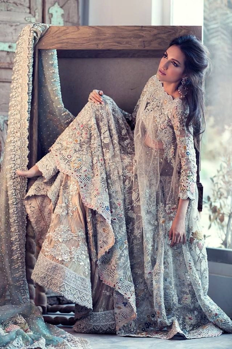 May include: An intricately designed, light-colored formal gown with elaborate embroidery and lace detailing. The gown features long sleeves and a flowing skirt, with colorful accents. A sheer, matching veil drapes over the shoulders.