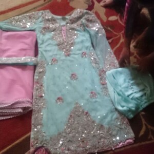May include: A light blue and pink outfit with silver embellishments. The outfit includes a long-sleeved top, a pair of trousers, and a dupatta.