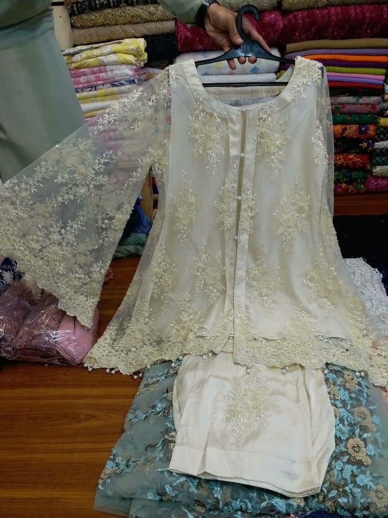 May include: A white, sheer, embroidered top with long sleeves and a button closure. The top is layered over a white, embroidered skirt. The fabric is delicate and has a lace-like texture.