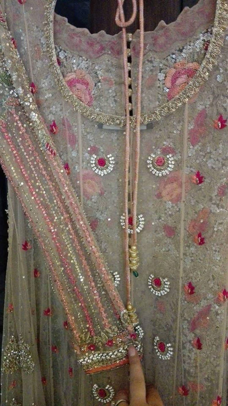 May include: A beige net fabric garment with pink and gold embroidery. The garment has a round neckline and a long, flowing design. The fabric is embellished with sequins and beads.