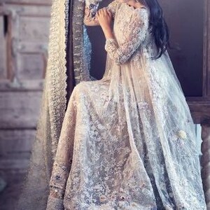 May include: A long, flowing, ivory-colored gown with intricate lace detailing. The dress features long sleeves and a full skirt, with delicate embroidery and embellishments. A matching shawl is draped over a wooden chair.