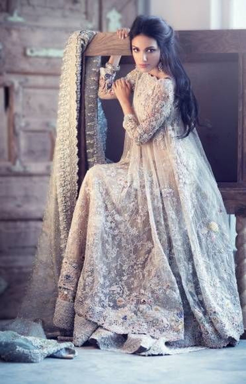 May include: A long, flowing, ivory-colored gown with intricate lace detailing. The dress features long sleeves and a full skirt, with delicate embroidery and embellishments. A matching shawl is draped over a wooden chair.