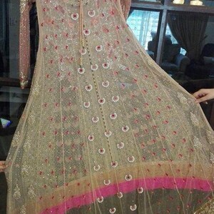 May include: A beige and pink net dress with intricate embroidery and sequins. The dress has a round neckline and long sleeves. The bottom of the dress has a pink and gold trim.