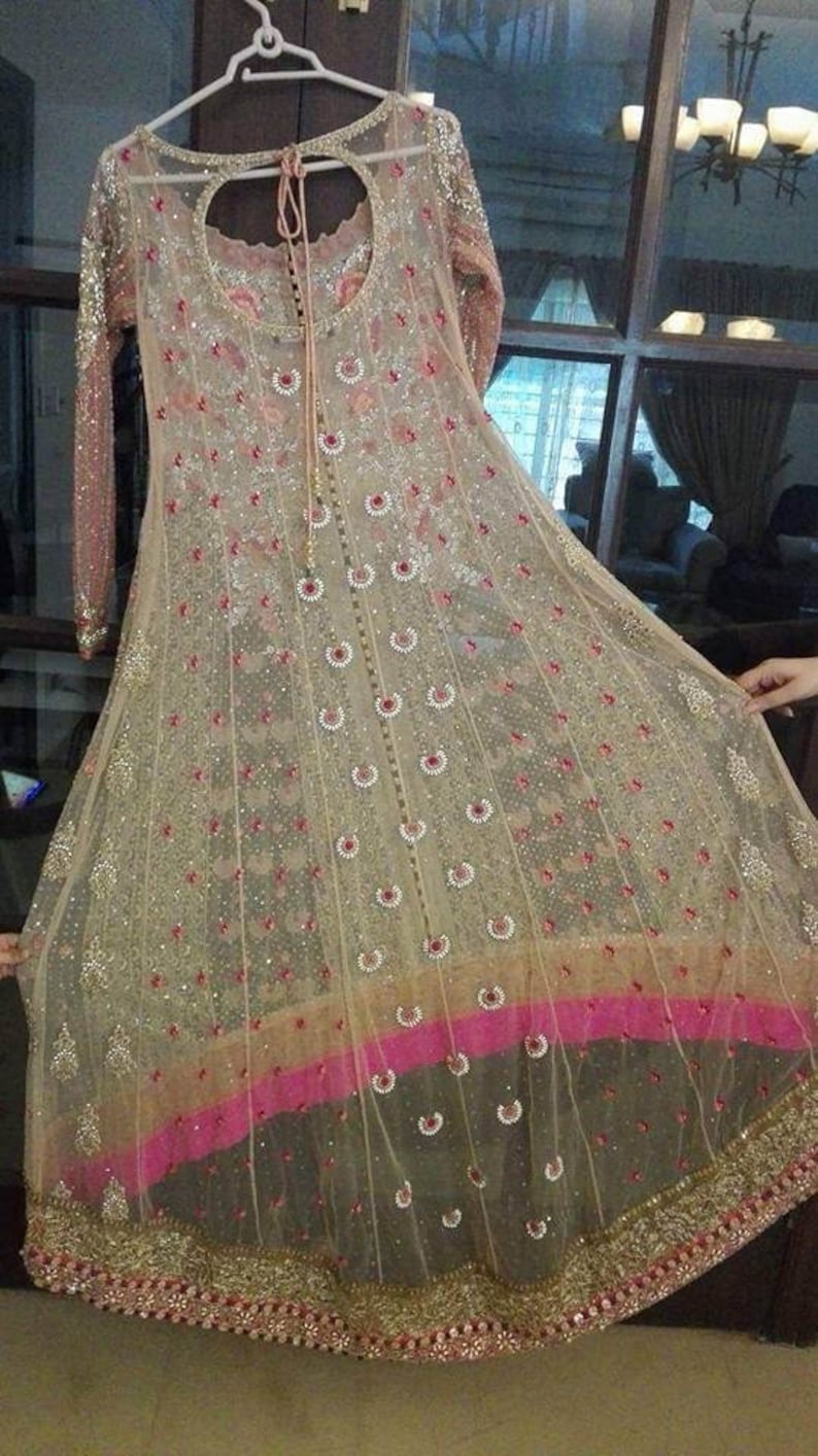 May include: A beige and pink net dress with intricate embroidery and sequins. The dress has a round neckline and long sleeves. The bottom of the dress has a pink and gold trim.