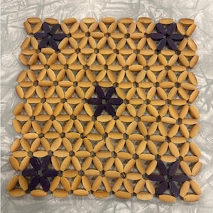 May include: A square wooden trivet with a repeating floral pattern. The trivet is made of small, light brown wooden pieces arranged in a geometric design. Dark purple wooden flowers are interspersed throughout the pattern.