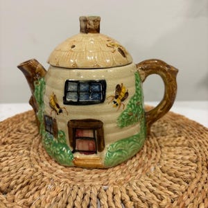 May include: A ceramic teapot shaped like a cottage with a thatched roof lid. The beige body features a brown door, a blue-paned window, green foliage, and painted bees. It has a brown branch-like handle and a matching spout, with a glossy finish.