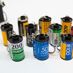 May include: A collection of film roll keychains in various colors, including yellow, black, blue, and red. Each keychain features film brand labels and a metal keyring. A vintage camera is visible in the background.